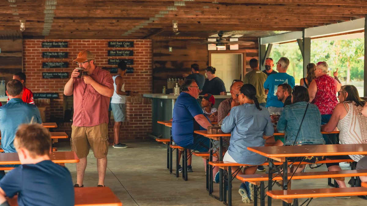 Inside the pavilion: a brick bar wall hung with chalkboard beer signs, a man sipping a pint, guests at orange beer-garden tables
