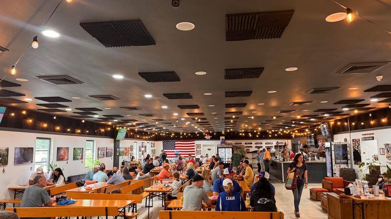 The taproom down its length: long tables full of people, string lights, a US flag on the far wall