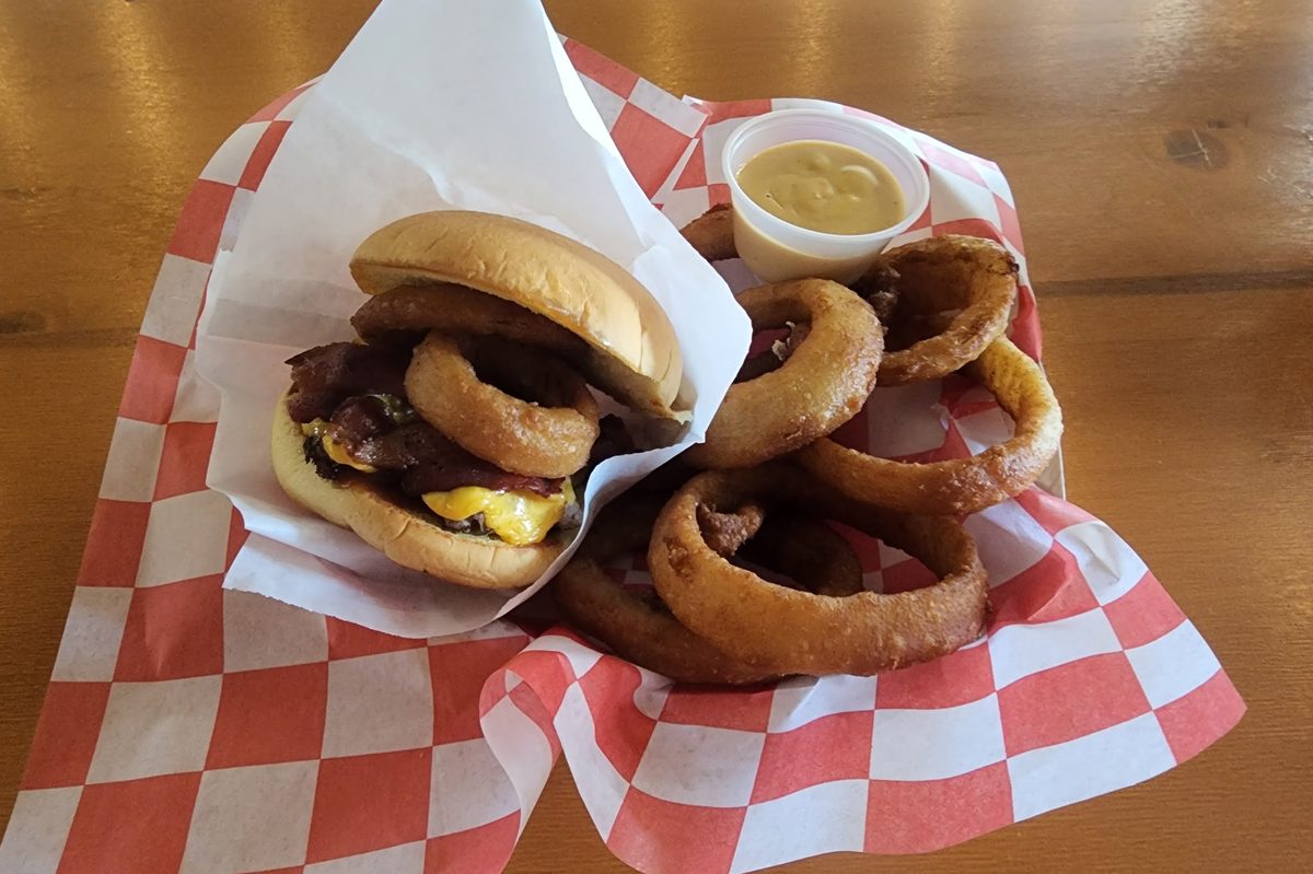 A bacon cheeseburger topped with onion rings, more rings and a cheese-sauce cup in red-checkered paper
