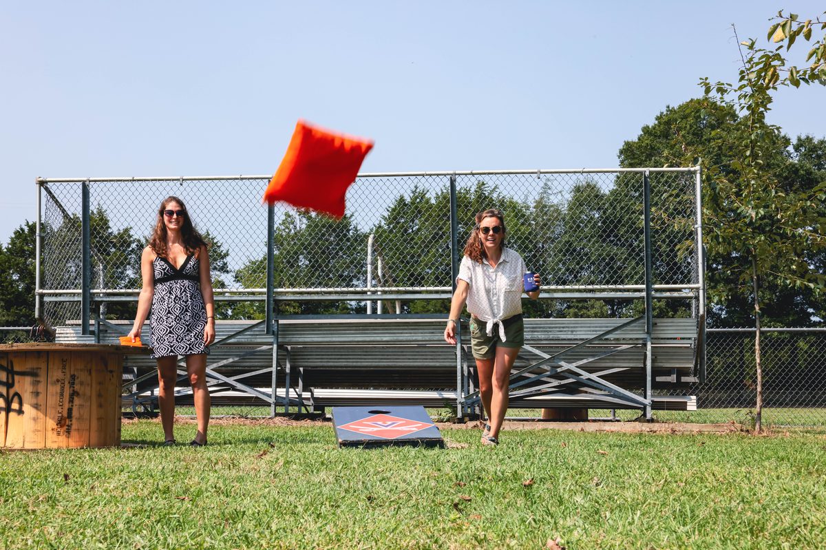 Two women playing cornhole on the lawn, a bag mid-air, the ball-field bleachers and backstop behind