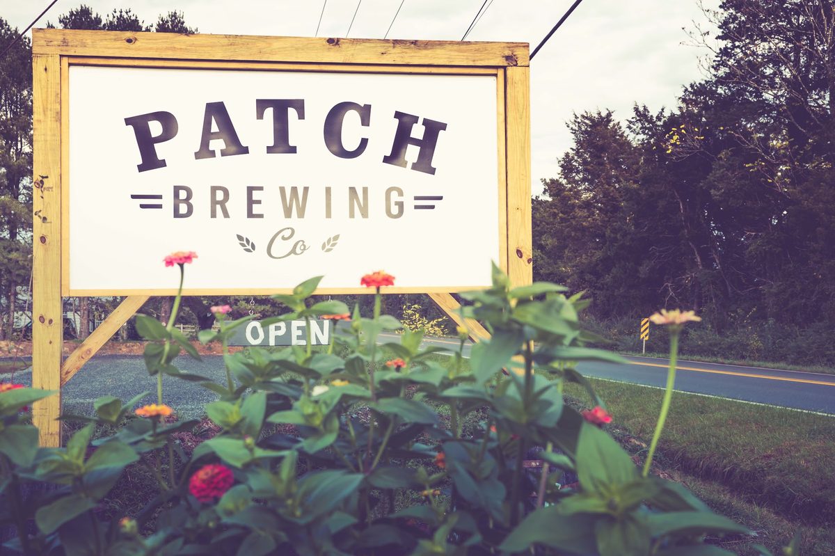 The roadside sign: PATCH BREWING Co on a timber frame with an OPEN plaque, zinnias in front