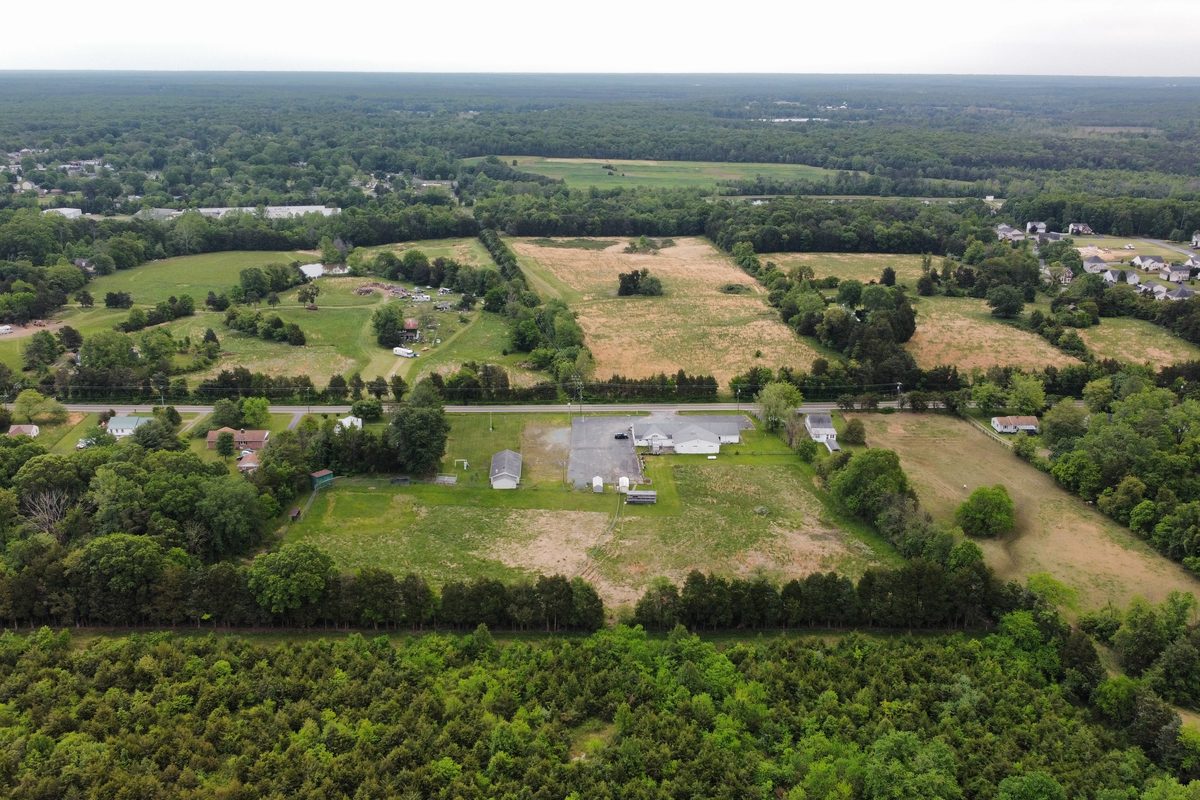The property from the air before the build: a white single-storey building and a paved lot beside the road, fields and woods around
